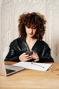 Positive retro businesswoman with curly hair sitting at table and using smartphone in office
