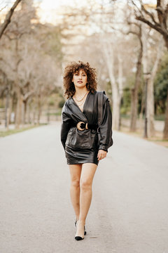 Optimistic female entrepreneur in trendy leather jacket and with curly hair smiling at camera while standing on blurred background of street