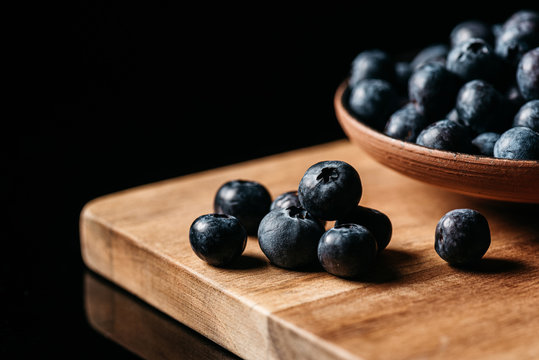 Fresh ripe blueberry placed on wooden table near bowl with berries against black background