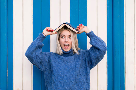 Funny astonished young female in casual knitted sweater holding open book on head and looking away while standing against colorful wall