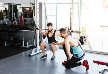 Powerful determined sportsman doing exercise with barbell at squat rack during weightlifting training with personal coach in modern gym