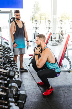 High angle of calm confident adult man in activewear performing single leg squat exercise with dumbbell while leaning on bench against blurred interior and sports equipment in modern spacious fitness club