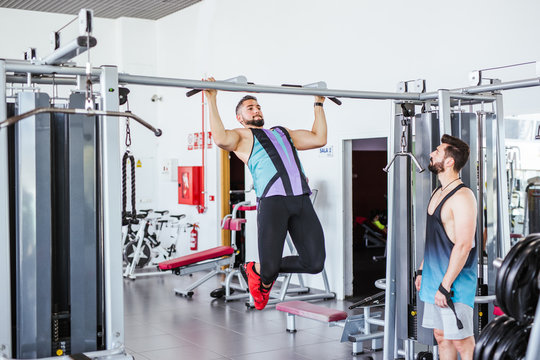 Low angle of pensive instructor watching determined athlete in sportswear performing pull ups exercise on bar in contemporary sport club