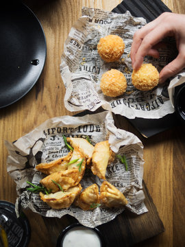 From above top view cropped unrecognizable person hand holding delicious deep fried cheese balls near plate with dumplings garnished with green herbs placed on paper served with white sauce on wooden table