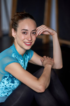 Happy professional ballet dancer sitting on the floor looking at camera resting after dancing training on a modern dark academy studio