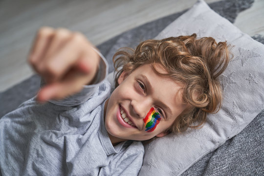 Cheerful boy with rainbow under eye smiling and pointing at camera while lying on blanket and pillow at home during quarantine