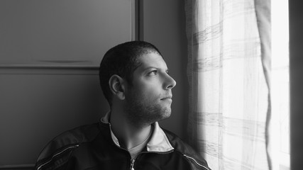 Black and white headshot of a short hair and short beard young man looking sad at the window