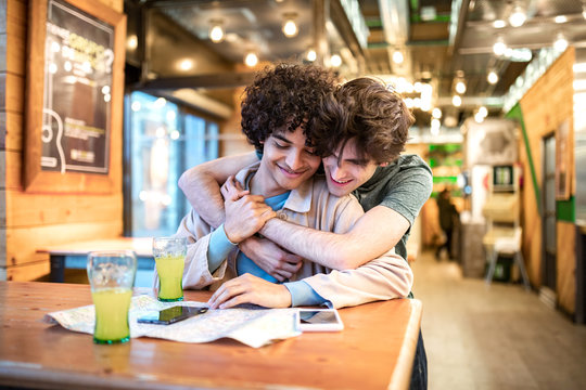 Multiethnic young homosexual men embracing each other looking at direction navigation map and having fresh drinks smiling while sitting at cafe table during romantic date