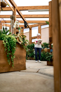 Full body elderly gardener writing on clipboard while standing near plants in wooden greenhouse