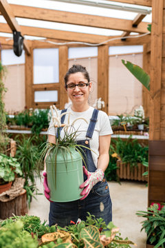 Happy adult woman smiling to the camera and carrying can with blooming flower while working in terrace garden