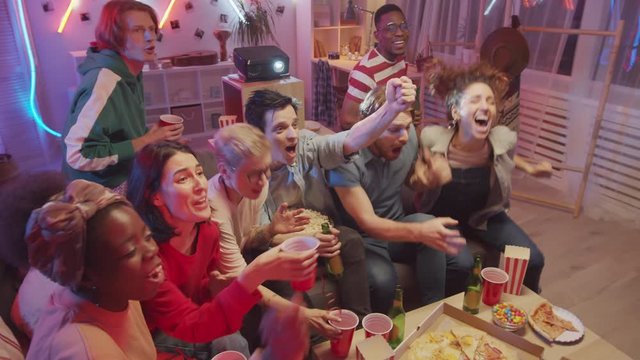 High angle view of group of young joyous friends watching sports match on TV while sitting together on couch, then yelling in excitement and toasting with beer while celebrating goal