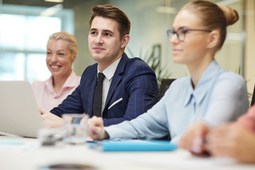 Young businessman in suit sitting at the table and listening to his colleagues during teamwork at meeting at office