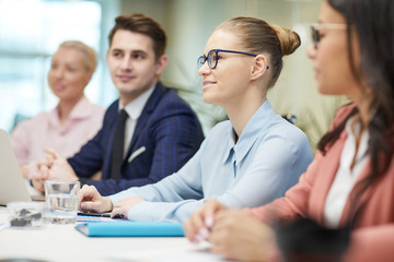 Fototapeta premium Young smiling businesswoman in eyeglasses sitting at the table and talking to her colleagues during business meeting at office