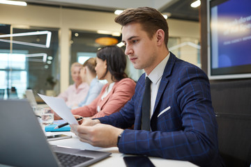 Young businessman sitting at the table in front of laptop and reading a document with his colleagues working in the background