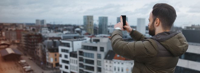 Back view of young man on the roof, taking photo of city by smartphone.