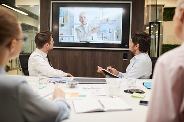 Rear view of group of business people sitting at the table and listening to mature businesswoman on the screen during online business conference at office