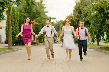 Newlyweds are have fun at wedding day together with their friends, holding hands they run along the street.