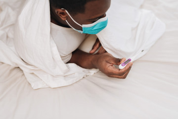 African American male in blue and white surgical mask laying in bed looking at a thermometer, displaying signs of being sick