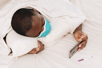African American man laying in bed with blue and white surgical face mask looking at cellphone, displaying signs of sickness