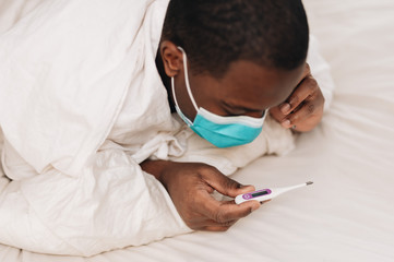 African American male in blue and white surgical mask laying in bed looking at a thermometer, displaying signs of being sick