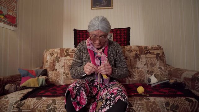 Soviet-style granny calmly knits while watching TV &mdash; parody of traditional babushka in retro apartment with serious face and comic timing