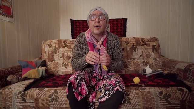 PARODIC SOVIET-STYLE GRANNY knits and stares at TV with deadpan face &mdash; comic retro look, cheap sofa, old interior, exaggerated character performance