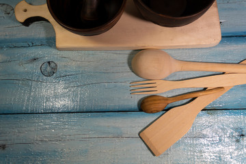 On a wooden background are wooden cutlery - a spatula, two spoons, a fork. Two clay bowls are visible on a cutting board.