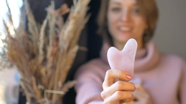 The girl shows a massage gua sha massager made from natural stone rose quartz that is used to stimulate lymphatic system and has anti age effect, female face and spikelets are not in focus