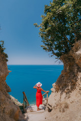 girl in a red dress and a white hat stands on the steps against the sea