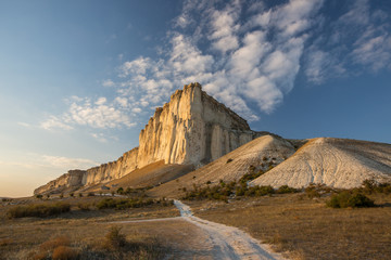Obraz premium White rock against a beautiful sunset, light clouds fly over the rock and the road leads to the foot of the rock.