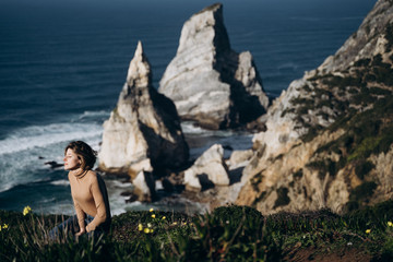 Naklejka na ściany i meble Young woman is resting and meditating near Praia da Ursa Beach Rocks on sunset. Sintra, Portugal. Atlantic Ocean coast
