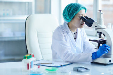 Female doctor sitting at the table and examining blood samples using microscope in her work at the...