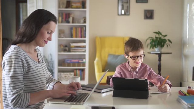 Young mother is working with laptop and son doing homework at table in home interior spbd. Beautiful american woman uses computer for work while child writes in notebook, sitting at desk in room