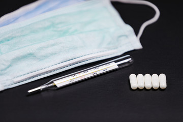 Einwegmundschutzmaske mit einem Quecksilber Fieberthermometer und Tabletten auf einer schwarzen Schieferplatte.