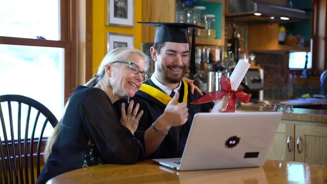 In front of a laptop computer in a home dining room, a mother and her son in graduation attire talk and wave to someone they are teleconferencing with.