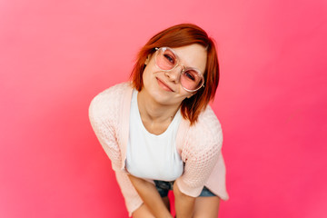 Pretty young redhead woman wearing glasses leaning forwards with a cute smile as she stares at the camera isolated on pink background