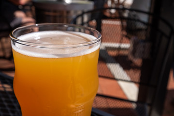 Beer in a clear glass with a small froth. The glass is on a patio table at a microbrewery. The sour beer has an orange color to the glass. There's sun shining on the ground in the background.  