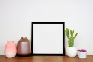 Mock up black square frame with home decor and potted plants. Wooden shelf against a white wall. Copy space.