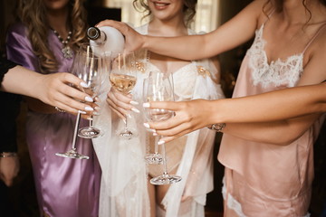 Female hands holding glasses of champagne at hen party. Bride and bridesmaids toasting with champagne and having fun on the wedding morning. Close up photo of girls celebrating a bachelorette party.