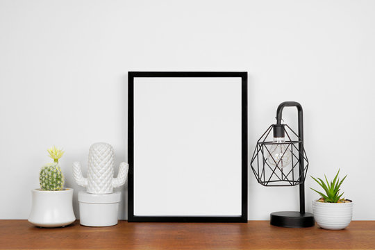 Mock up black portrait frame with home decor and potted plants. Wooden shelf against a white wall. Copy space.