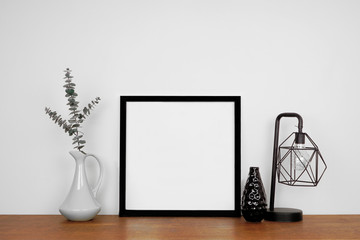 Mock up black square frame with home decor. Wooden shelf against a white wall. Copy space.