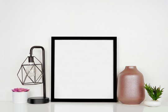 Mock up black square frame with home decor and potted plants. White shelf against a white wall. Copy space.