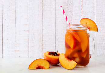 Refreshing peach iced tea in a mason jar glass. Side view with cut fruit against a white wood...