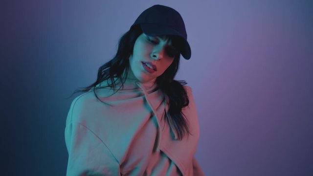 Trandy woman in cap dancing in Multi-Colours Neon Lighting room at night. Portrait of Young Stylish Woman Dancing at Party in nightclub
