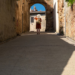 Fototapeta na wymiar a young woman with hat and traveler backpack on a narrow street with an arch on a sunny summer day in Tuscany, Italy