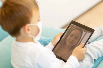Fototapeta premium Close-up of cute son with medical mask. sitting on sofa and using tablet while chatting with mom online.Visible mom face.During Covid-19 pandemic isolation.Stay home