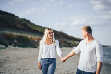 Happy young couple having fun at beach on sunny evening. Smiling teens on vacation. Laughing couple in love holding hand enjoying summertime. Guy leading his girlfriend