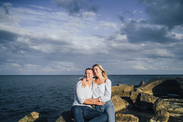 Two lovers blondes spending a romantic evening on the beach. They  feeling evening sun, hugging, kissing and laughing, sitting on the worm stones. The girl is sitting on the arms of a guy.