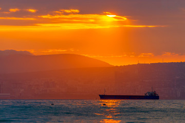 Naklejka na ściany i meble Cargo ship sailing the sea at a golden sunset or sunrise. Ocean shipment across water as the sun sets or rises. Harmony and beauty in nature. Scenic peaceful sea view