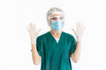 Portrait of a nurse girl on a white background in a green dressing gown, a protective mask, white medical cap and glasses. Woman look at the camera and holds hands in gloves raised up. Copy space.